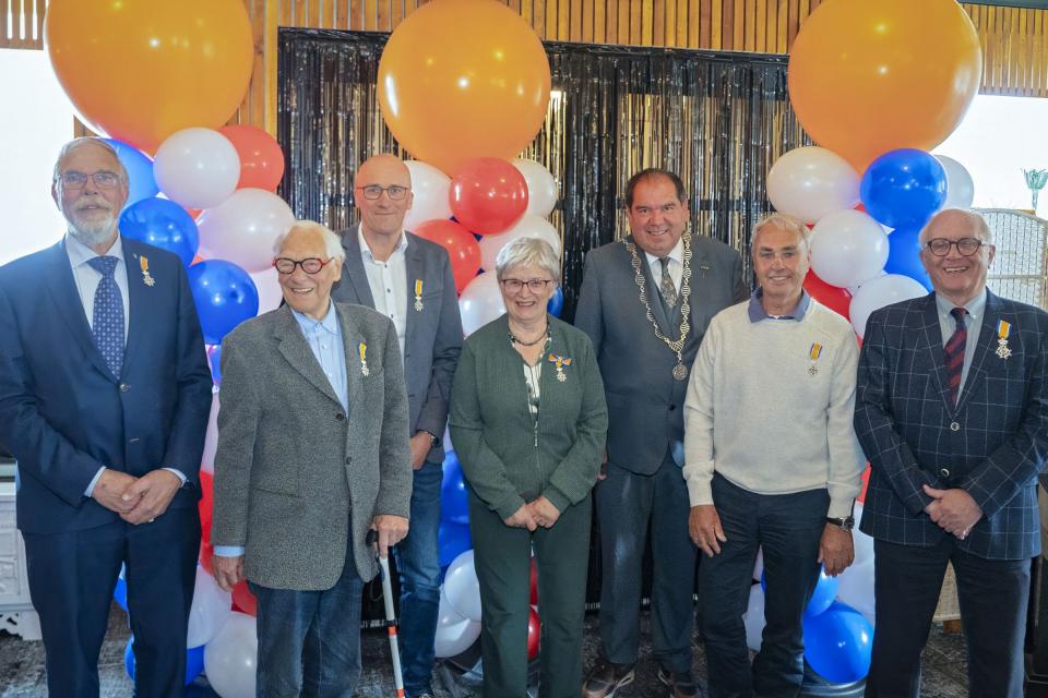 Zes gedecoreerden poseren trots met hun lintje en de burgemeester kijkt toe